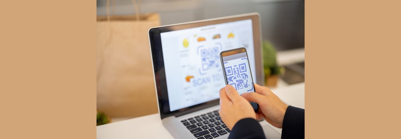 Person scanning a QR code with a smartphone in front of a laptop, illustrating the risk of online scams and digital fraud.